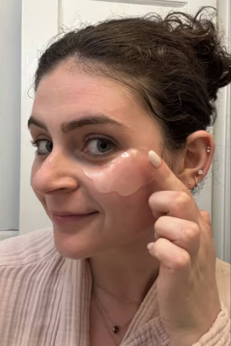 Woman smiles while applying translucent curved facial patches to her cheek during skincare routine.