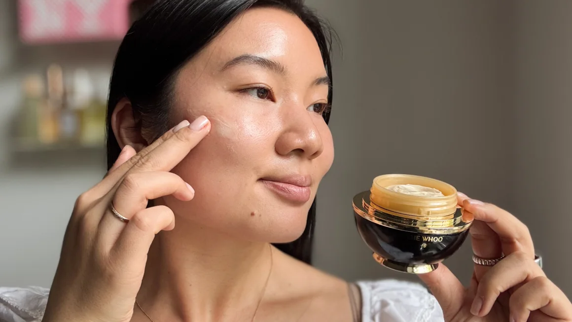 Woman applies moisturizer to her cheek from a jar of skincare product held in her other hand.