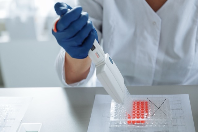 Lab technician wearing blue gloves uses a pipette to dispense liquid into a 96-well plate with orange wells at a lab bench, X marked on one area.
