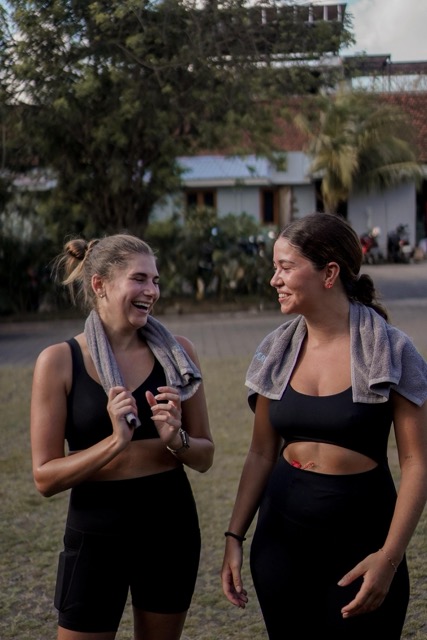 Two young women in black athletic outfits laugh together outdoors, towels draped over their shoulders near a building and trees.