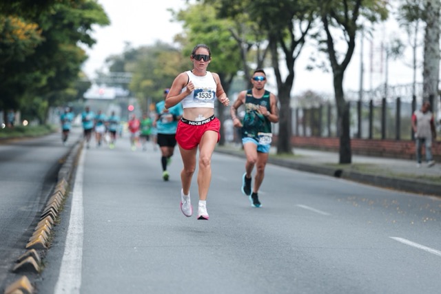 Female runner wearing a white tank top and red shorts leads a road race on a tree-lined street, bib number 1535.
