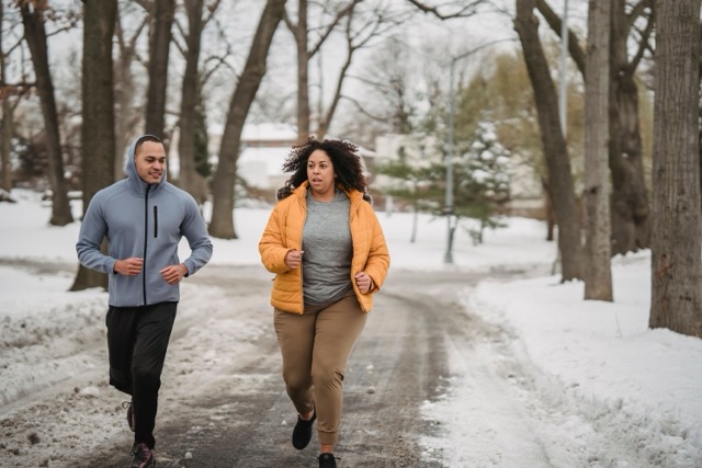 Two people jogging along a snow-covered park path on a winter day, wearing warm jackets.