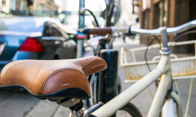 Close-up of a brown leather bicycle saddle on a cream bike, with a city street in the background.