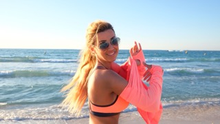 Smiling woman in an orange bikini top and pink cover-up on a sunny beach, with waves at the shore in the background.